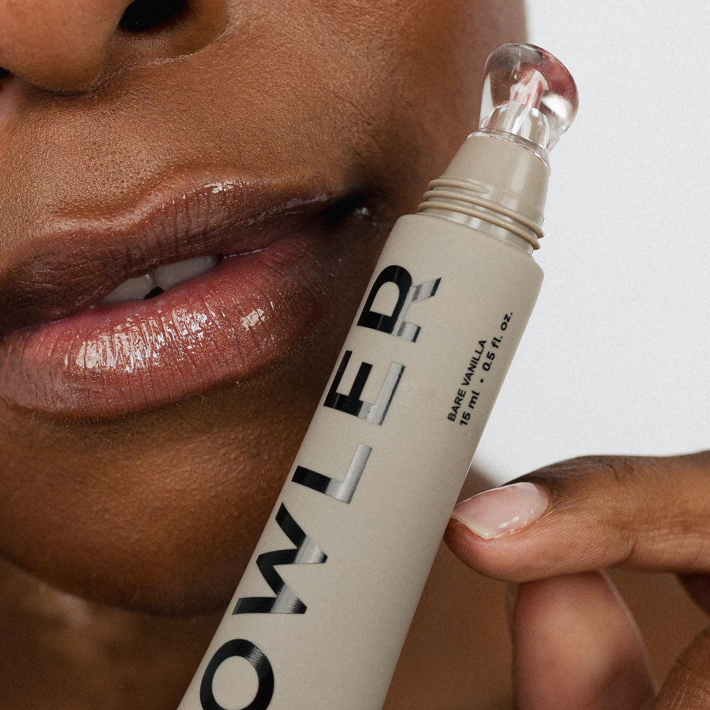 A close-up of a person holding a tube of Bowler Lip Gloss Treatment against their lower face, showing the beige tube with black lettering and a clear applicator tip.